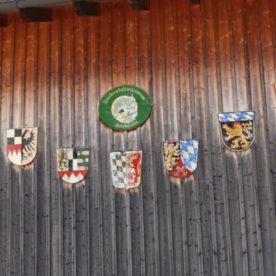 Das Logo vom Trachtenkulturzentrums Holzhausen mit den Wappen der Regierungsbezirke Bayerns auf einer Holzwand