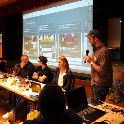 Matthias Penzenstadler gibt mit einem Mikrofon in der Hand vor einer Leinwand auf der Fotos abgebildet sind Einweisungen zum Fake or Real Workshop