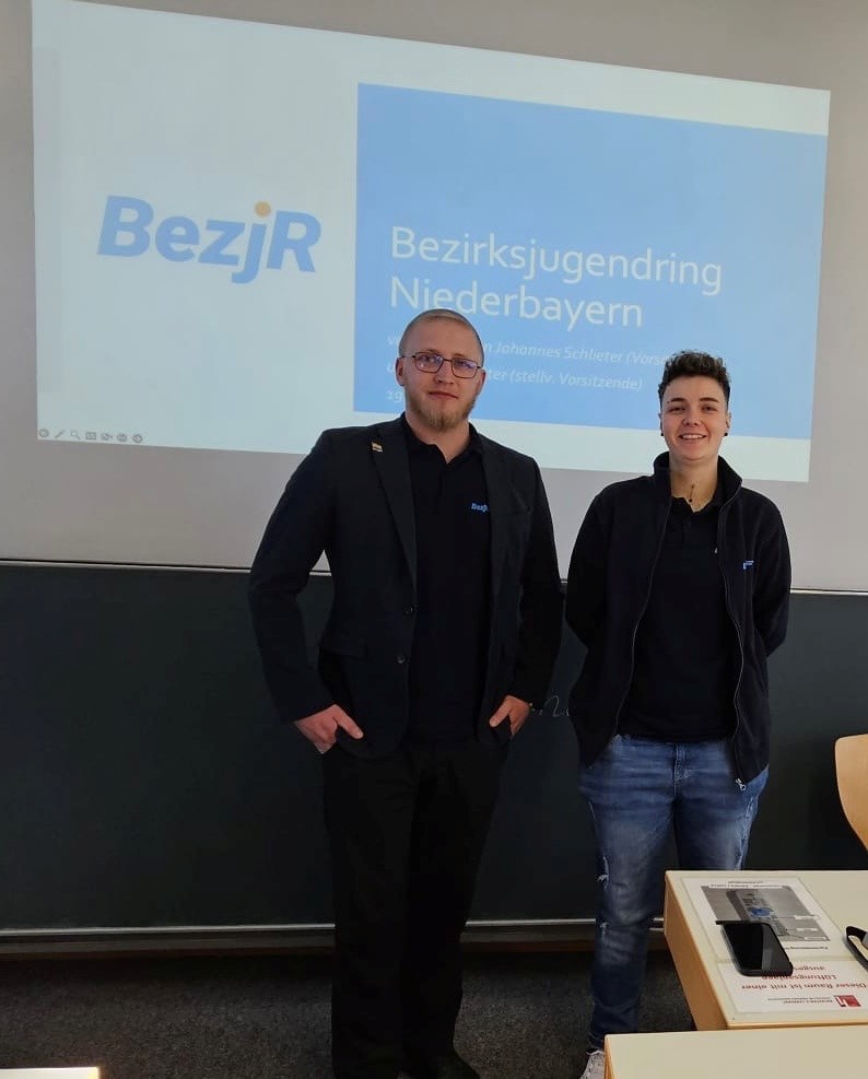 Vorsitzender Johannes Schlieter und stellvertretende Vorsitzende des Bezirkjugendring Niederbayern vor einer Leinwand auf der das Logo BezJR zu sehen ist.