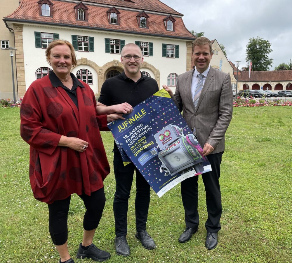 Anne Wachter, Johannes Schlieter und Bezirkstagspräsident Dr. Olaf Heinrich mit dem JUFINALE-Plakat vor einem Gebäude der Bezirksklinik Mainkofen