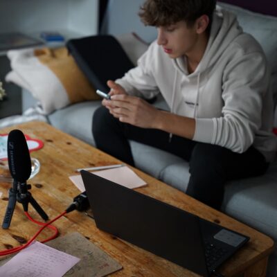 Ein Teilnehmer sitzt auf einer Couch mit Smartphone, Laptop und Aufnahmemikrophon.